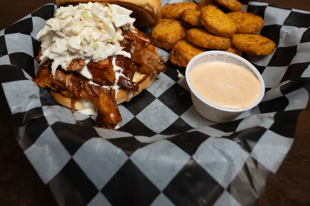 A pulled pork sandwich topped with coleslaw, served with tots and a dipping sauce at 212