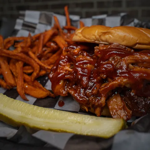 A pulled pork sandwich tossed in BBQ with sweet potato fries and a pickle